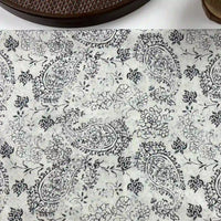 White surface features the Paisley Printed Voile Fabric by Longan Craft: pure cotton with black floral and paisley motifs. Part of a woven dark brown circular object appears at the top edge. Ideal for shirts, sold by the yard.