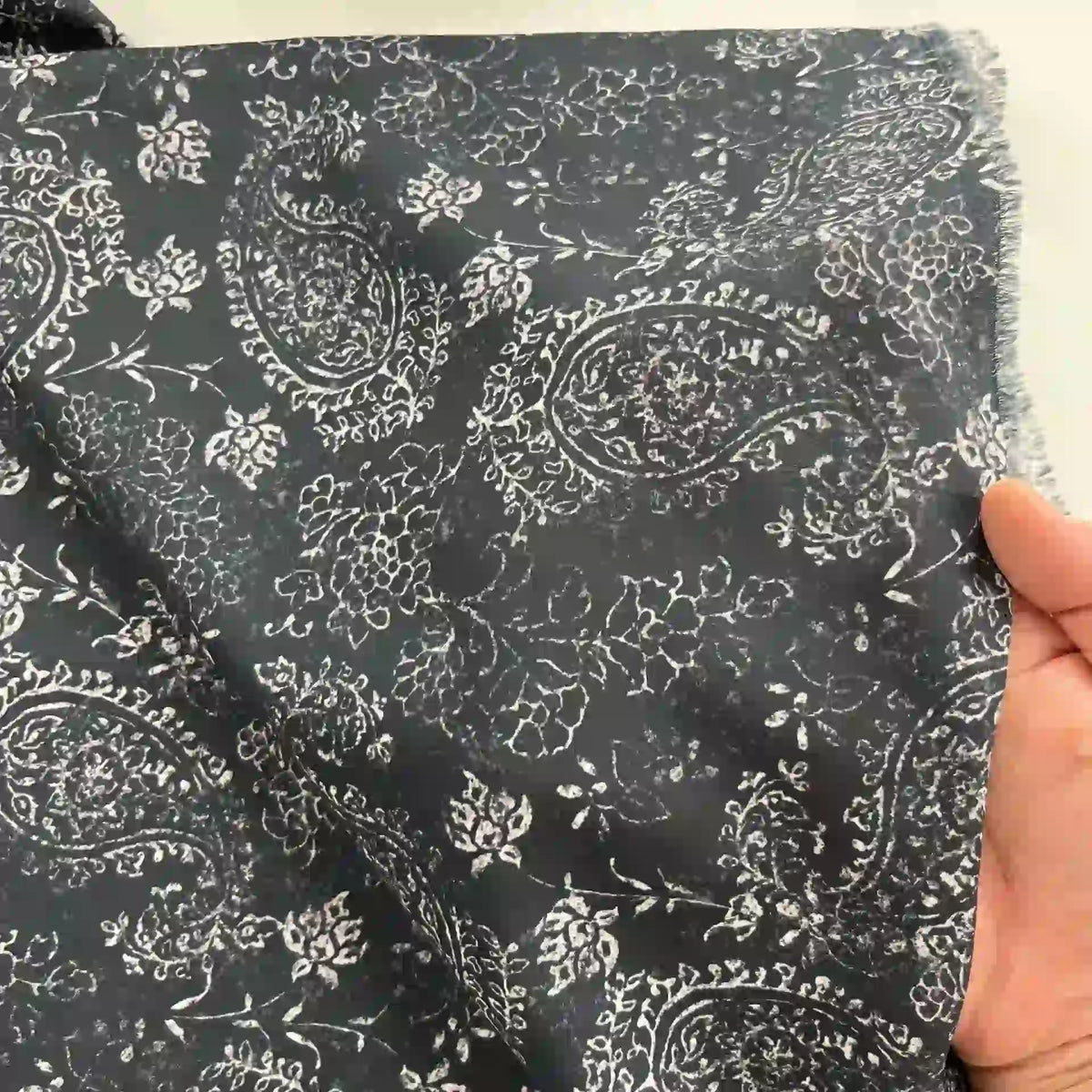 A hand holds Longan Craft's Paisley Printed Voile Fabric By The Yard, a pure cotton material in dark tones with a white floral pattern and slightly frayed edges for rustic charm—perfect for comfortable shirts.