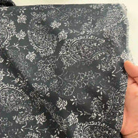 A hand holds Longan Craft's Paisley Printed Voile Fabric By The Yard, a pure cotton material in dark tones with a white floral pattern and slightly frayed edges for rustic charm—perfect for comfortable shirts.