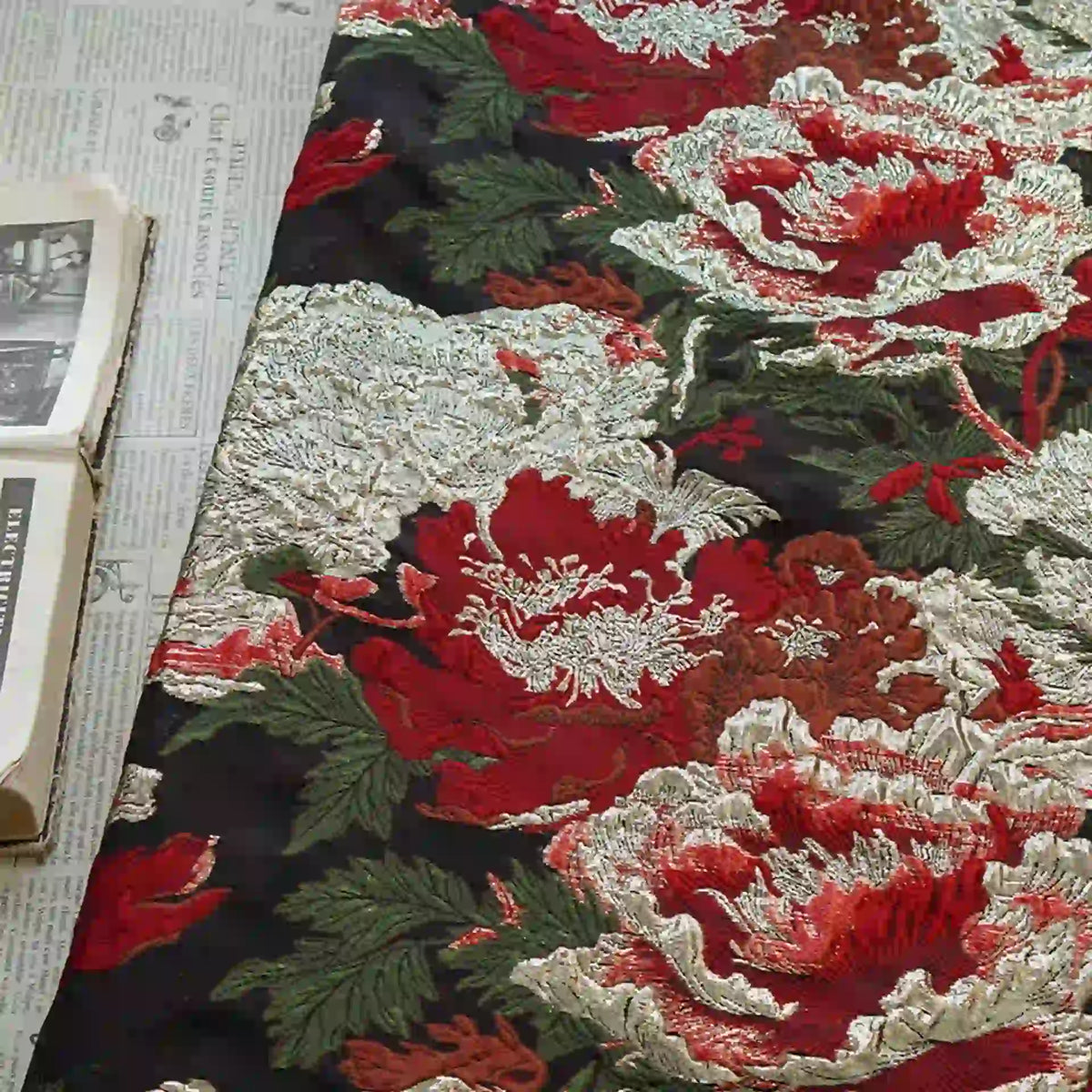 An open book rests on newspaper beside Longan Craft's Peony Embossed Metallic Brocade Fabric by the yard, a luxury golden black floral jacquard featuring large red, white, and pink peonies with green leaves on a dark background.