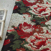 An open book rests on newspaper beside Longan Craft's Peony Embossed Metallic Brocade Fabric by the yard, a luxury golden black floral jacquard featuring large red, white, and pink peonies with green leaves on a dark background.
