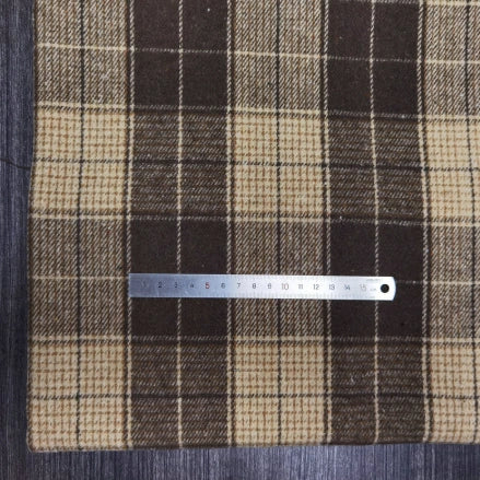 A Longan Craft Plaid Brushed Mammoth Flannel Fabric in brown and beige rests on a grey wooden surface, with a 20 cm metal ruler placed horizontally across the colorful polyester cotton material.