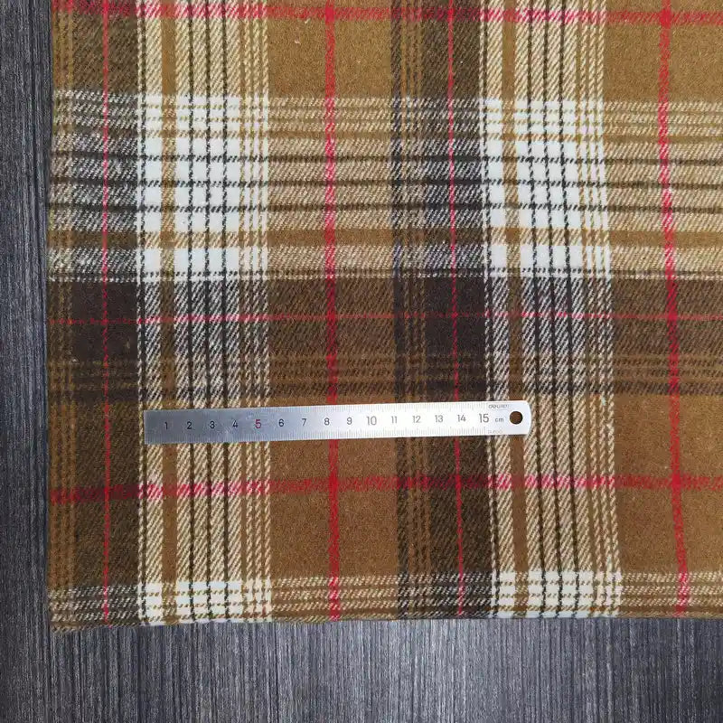 A metal ruler measures 0–15 cm on Longan Craft's Plaid Brushed Mammoth Flannel Fabric By The Yard—a comfy, colorful polyester cotton fabric with brown, tan, and red plaid—over a grey wood-textured surface.