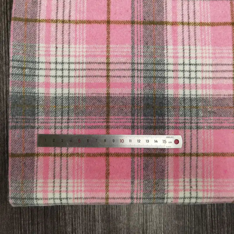 A Longan Craft Plaid Brushed Mammoth Flannel Fabric (polyester-cotton), shown in pink, white, gray, and brown plaid, is displayed on a dark wood surface with a 15 cm metal ruler measuring its width.