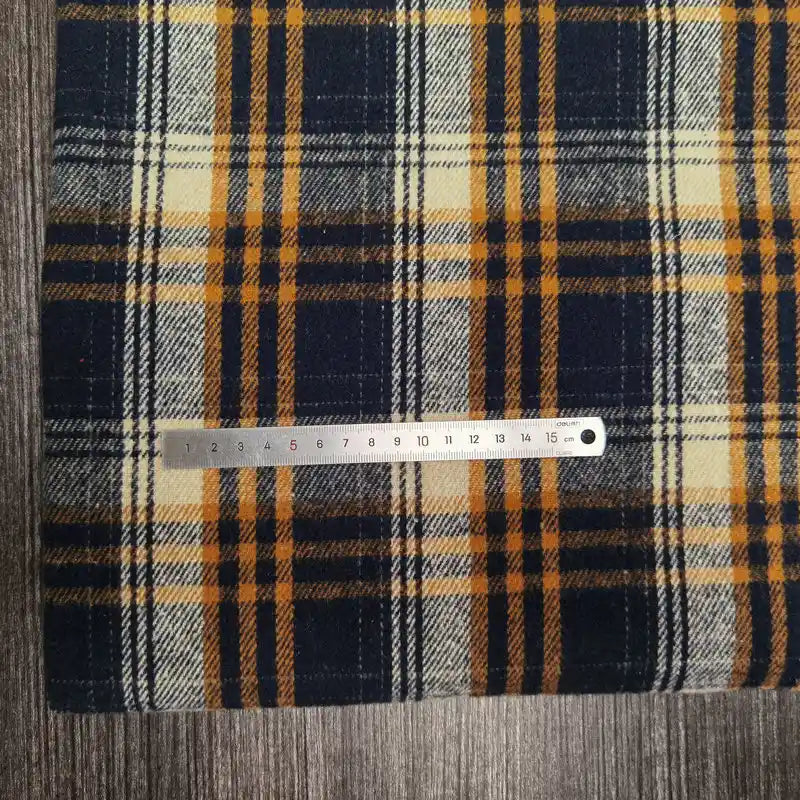 A metal ruler lies on top of Longan Craft's Plaid Brushed Mammoth Flannel Fabric By The Yard—colorful polyester cotton plaid flannel—resting on a grey textured surface. Ruler shows measurements in centimeters.