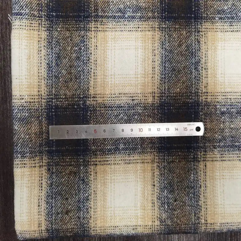 A metal ruler measuring 16 cm rests horizontally on Longan Craft's Plaid Brushed Mammoth Flannel Fabric, a colorful polyester cotton flannel with beige, navy blue, and brown checks, showing the scale of the pattern.
