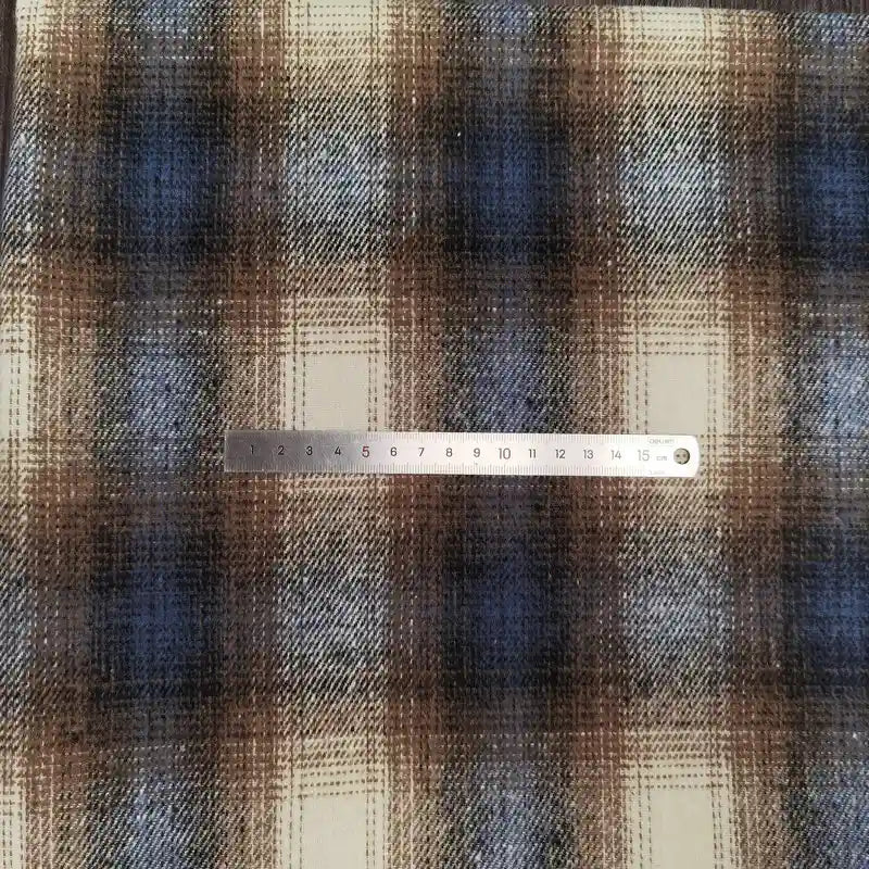 A 15cm metal ruler lies horizontally across Longan Craft's Plaid Brushed Mammoth Flannel Fabric, a comfy fall polyester-cotton material featuring a colorful brown, blue, and white plaid pattern.