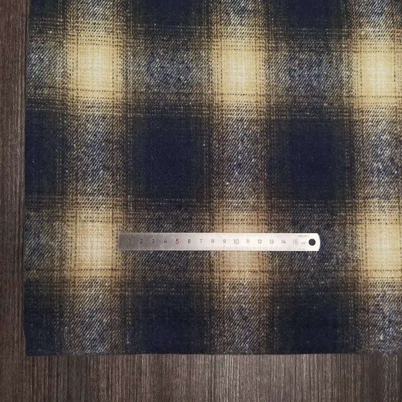 A steel ruler measures Longan Craft’s Plaid Brushed Mammoth Flannel Fabric by the yard, featuring a colorful polyester cotton pattern. The ruler lies horizontally across the comfy fall fabric on a dark wooden surface.