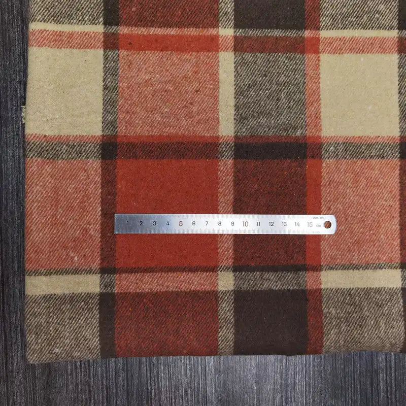 A metal ruler sits horizontally on Longan Craft’s Plaid Brushed Mammoth Flannel Fabric in red, orange, brown, and beige. The comfy polyester cotton material spans a dark wood surface; the ruler measures about 16 cm.