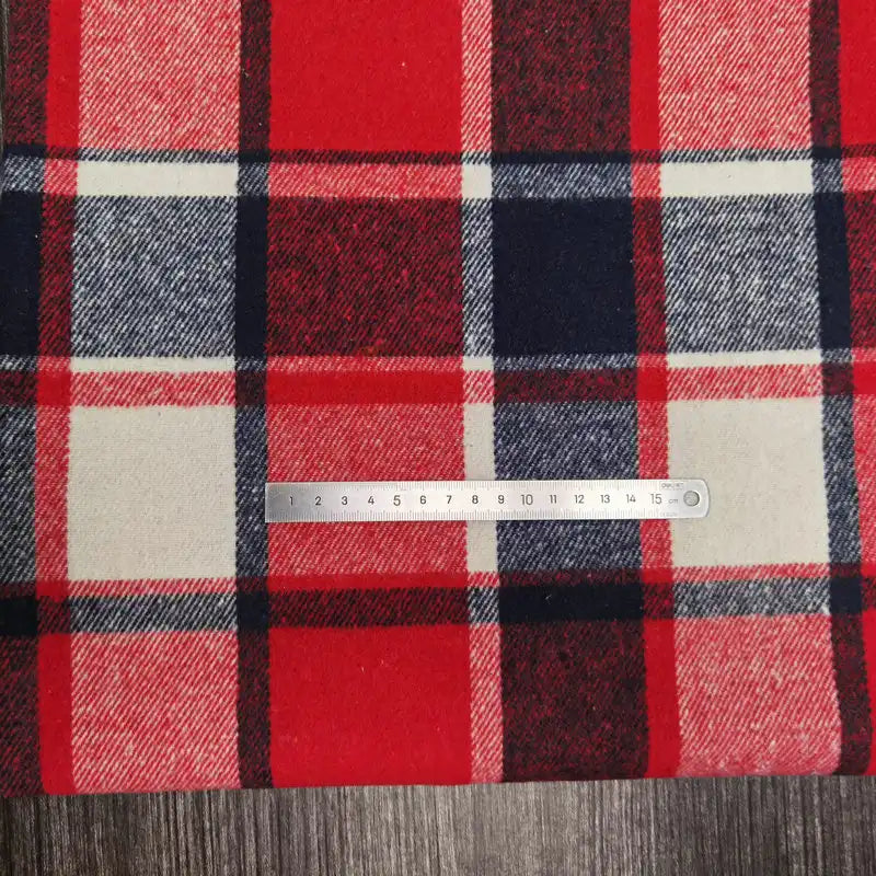 A Longan Craft Plaid Brushed Mammoth Flannel Fabric in red, navy, white, and black is shown on a dark wood surface with a metal ruler measuring one square; this cozy polyester cotton flannel is sold by the yard.
