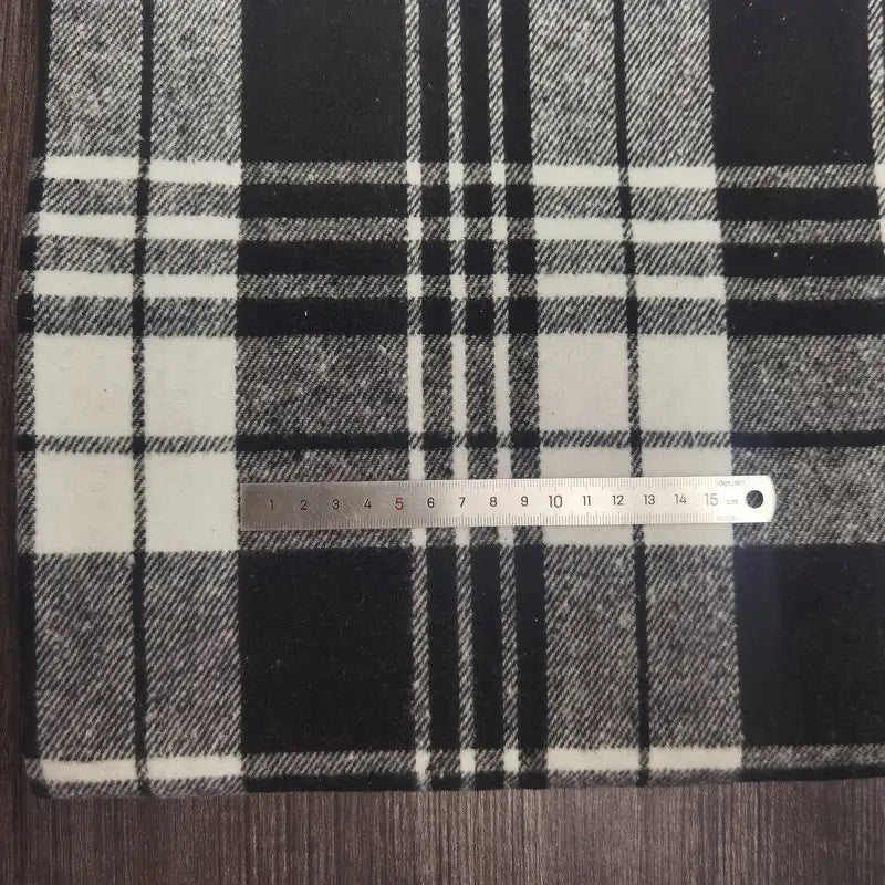 A metal ruler rests on Longan Craft's Plaid Brushed Mammoth Flannel Fabric By The Yard, a colorful polyester cotton flannel. The comfy fall fabric displays black, white, and gray plaid atop a wooden surface.