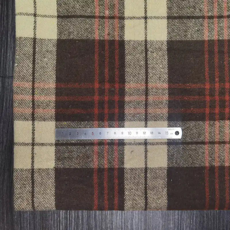 A metal ruler lies horizontally on Longan Craft's Plaid Brushed Mammoth Flannel Fabric by the Yard, a colorful polyester cotton material. The comfy fall flannel features brown, beige, and red stripes. The ruler displays centimeters on the cloth.