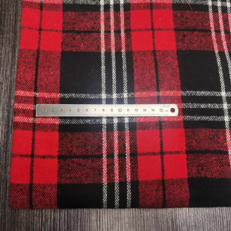 A silver ruler (16 cm) rests on Longan Craft's Plaid Brushed Mammoth Flannel Fabric By The Yard—a colorful polyester-cotton flannel—featuring a classic red, black, and white pattern atop a dark wooden surface.