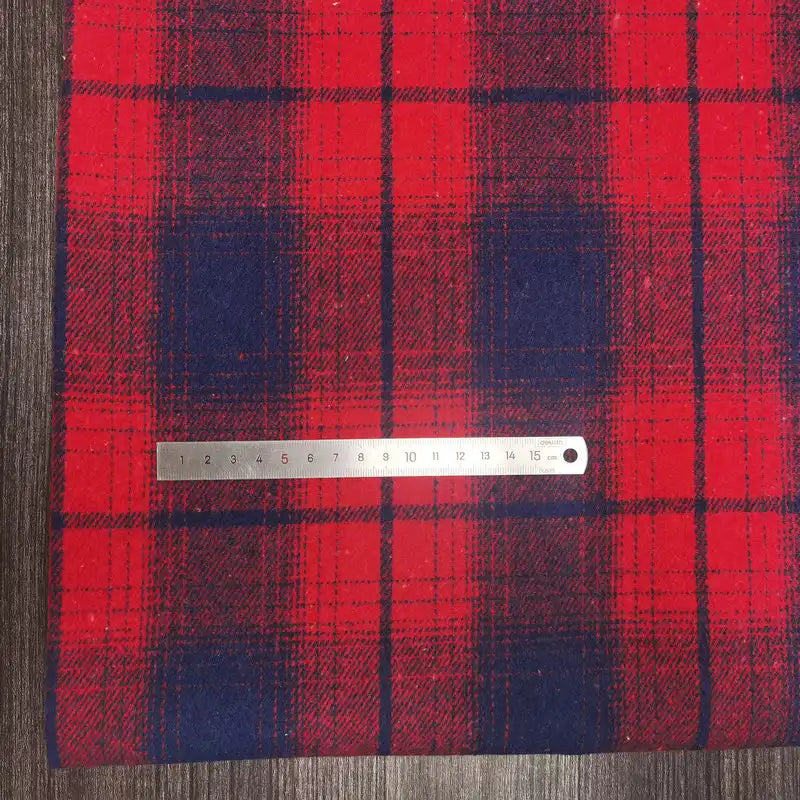 A silver ruler rests on Longan Craft Plaid Brushed Mammoth Flannel Fabric by the Yard, a comfy polyester-cotton material with large red and navy blue checks, displayed on a wood-textured surface. The ruler shows cm and mm measurements.