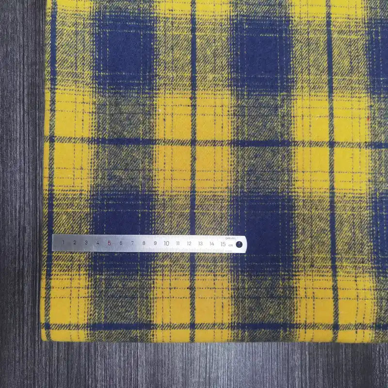 A Longan Craft Plaid Brushed Mammoth Flannel Fabric in yellow and blue is displayed on dark wood, with a metal ruler measuring up to 16 cm laid horizontally across the colorful polyester cotton material.