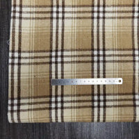 A metal ruler rests on Longan Craft's Plaid Brushed Mammoth Flannel Fabric By The Yard, a colorful polyester cotton flannel, in brown, beige, and white plaid, set on a dark wooden surface with the ruler measuring about 18 centimeters.