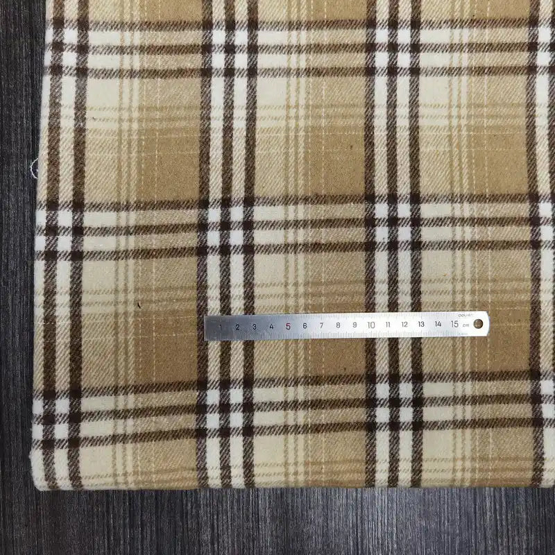 A metal ruler rests on Longan Craft's Plaid Brushed Mammoth Flannel Fabric By The Yard, a colorful polyester cotton flannel, in brown, beige, and white plaid, set on a dark wooden surface with the ruler measuring about 18 centimeters.