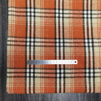 A Longan Craft metal ruler lies on Plaid Brushed Mammoth Flannel Fabric by the Yard, a comfy polyester-cotton material with orange, beige, black, and white stripes, all displayed on a dark wood surface. The ruler measures about 16 cm.