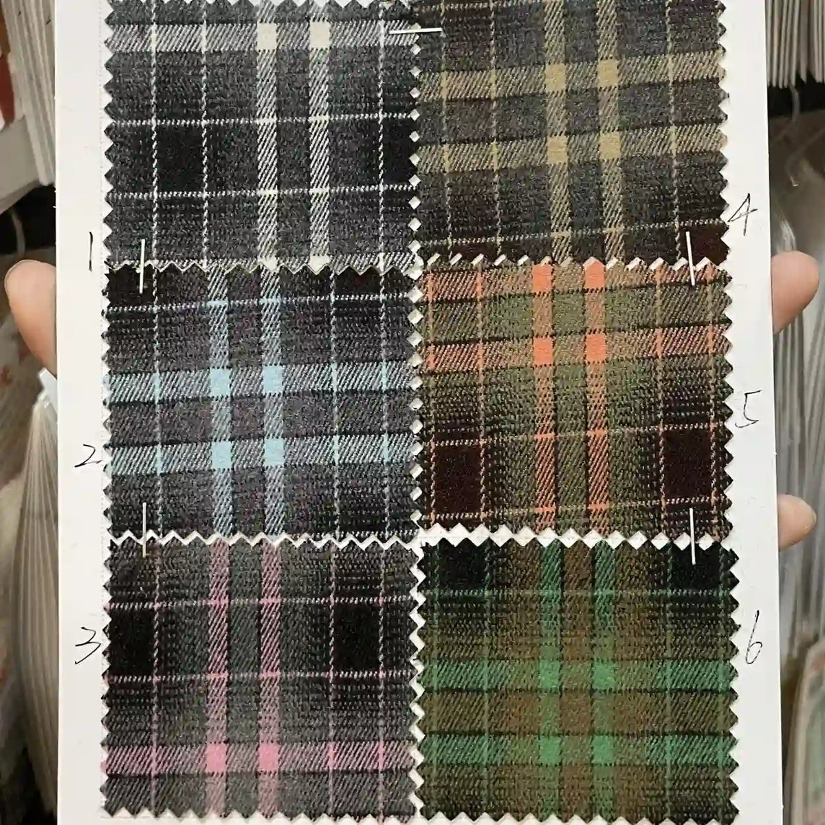 A hand holds a sheet showing six numbered swatches of Longan Craft's Plaid Mammoth Flannel Fabric By The Yard, each with unique plaid patterns on dark backgrounds. This comfy polyester rayon textile is ideal for shirting and crafts.