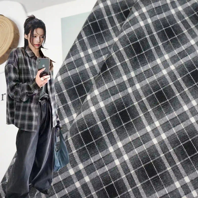 A woman wears a shirt made from Longan Craft's Plaid Mammoth Flannel Fabric By The Yard—comfy polyester rayon shirting flannel—in black and gray plaid, paired with loose black pants, taking a mirror selfie with a close-up of the same plaid pattern in the background.