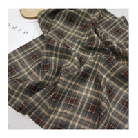 A piece of Longan Craft's Plaid Mammoth Flannel Fabric by the yard, a soft polyester rayon shirting material in beige, brown, green, and red plaid, is shown on a light surface with partial text in the corner.