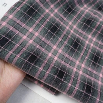 A hand holds a piece of Longan Craft Plaid Mammoth Flannel Fabric By The Yard in pink and dark green. Made from comfy polyester rayon, this shirting flannel textile is shown against a light background with partial printed text.