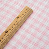 A wooden ruler with centimeter markings lies diagonally on the Longan Craft Plaid Print Double Gauze Fabric By The Yard, a pure cotton checked printed gauze material, highlighting the soft and breathable fabric ideal for cozy baby clothing.