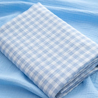 The Plaid Print Double Gauze Fabric by Longan Craft, featuring a blue and white checked cotton design, is neatly folded on top of a solid light blue textured cloth.