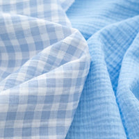 The Plaid Print Double Gauze Fabric by the yard from Longan Craft, made of pure cotton with a light blue and white checked design, is displayed alongside a solid light blue textured cotton fabric, both arranged in soft folds to highlight their breathable and cozy qualities—perfect for baby clothing.