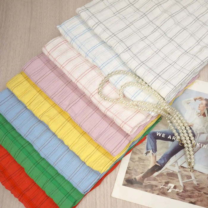 A stack of vibrant Longan Craft Plaid Rayon Seersucker Fabric By The Yard in red, green, yellow, purple, pink, blue, and white rests on a light wood surface beside a pearl necklace and fashion magazine. Wrinkle-resistant and breathable.