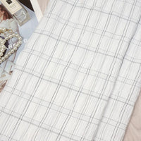 A neatly folded Plaid Rayon Seersucker Fabric by Longan Craft, featuring a subtle black grid, rests on a surface with pearls, a fashion magazine, and perfume. This breathable polyester textile is ideal for elegant dresses and décor.