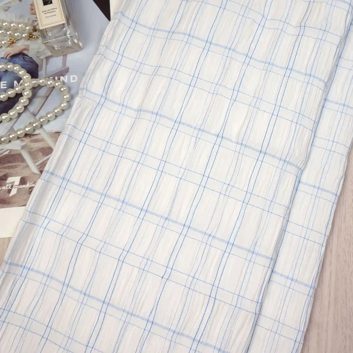 A Longan Craft Plaid Rayon Seersucker Fabric By The Yard with light blue checks is laid flat on a table, accompanied by a string of pearls, perfume bottle, and a magazine partially visible underneath.