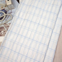 A Longan Craft Plaid Rayon Seersucker Fabric By The Yard with light blue checks is laid flat on a table, accompanied by a string of pearls, perfume bottle, and a magazine partially visible underneath.