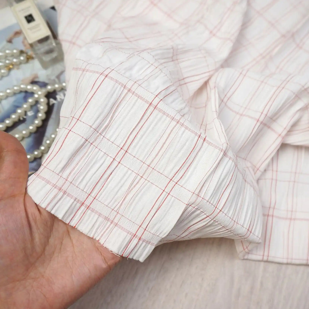 A hand holds the cuff of a white shirt with red plaid lines, made from Longan Craft’s Plaid Rayon Seersucker Fabric By The Yard—a breathable polyester seersucker textile ideal for dresses. Pearl necklaces, perfume, and fabric appear in the background.