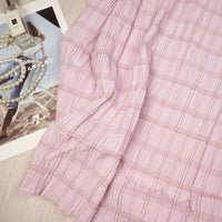 The Longan Craft Plaid Rayon Seersucker Fabric, a light pink checked polyester material ideal for dresses, is displayed draped on a flat surface beside a magazine, perfume bottle, and white pearls.
