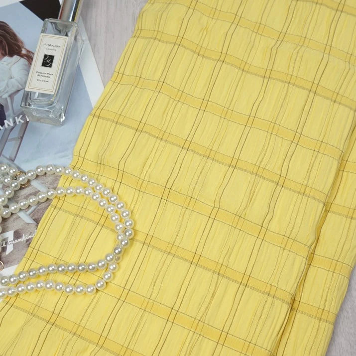 A piece of Longan Craft’s Plaid Rayon Seersucker Fabric by the yard is folded on a magazine, with Jo Malone perfume and white pearls beside it, showcasing the lightweight checked polyester textile ideal for dresses.