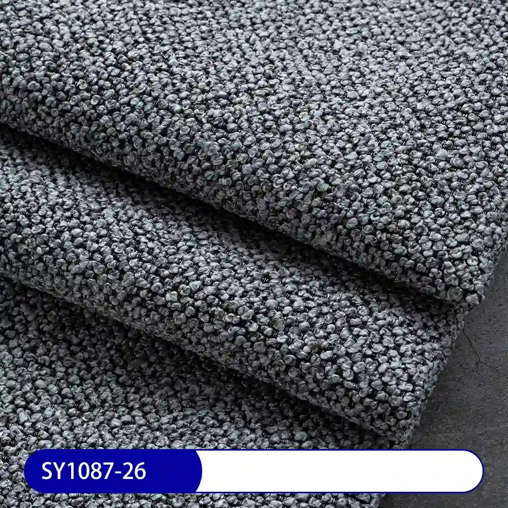 Close-up of folded, textured gray Plain Color Boucle Fabric By The Yard from Longan Craft, featuring a pebbled thick polyester surface for drapery or upholstery. A blue and white label at the bottom shows code SY1087-26.