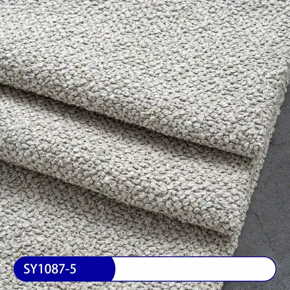 Three pieces of light gray Plain Color Boucle Fabric By The Yard from Longan Craft are stacked on a dark surface, with a blue and white label reading SY1087-5, showcasing this thick polyester boucle upholstery fabric.