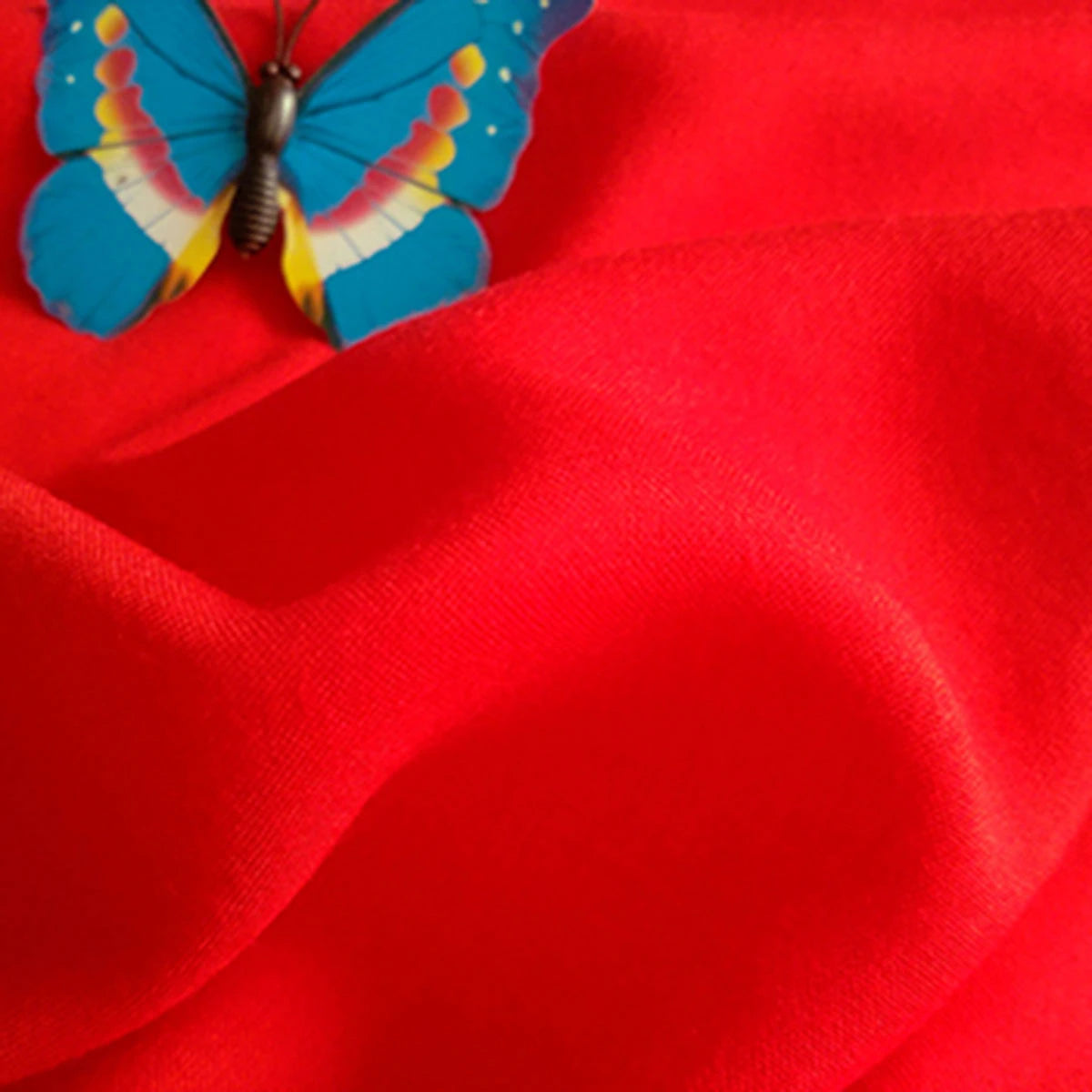 A vibrant blue butterfly with yellow and red markings sits on Longan Craft's bright red Plain Color Jersey Viscose Fabric—100% rayon viscose, perfect for dresses.