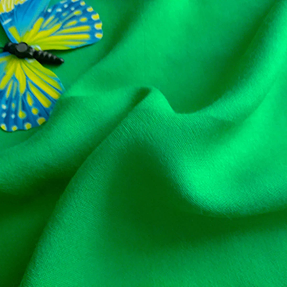 A close-up of Longan Craft’s Plain Color Jersey Viscose Fabric by the yard, 100% rayon viscose with a smooth texture, shown in vibrant green with a yellow and blue butterfly resting on its breathable surface—ideal for dresses.