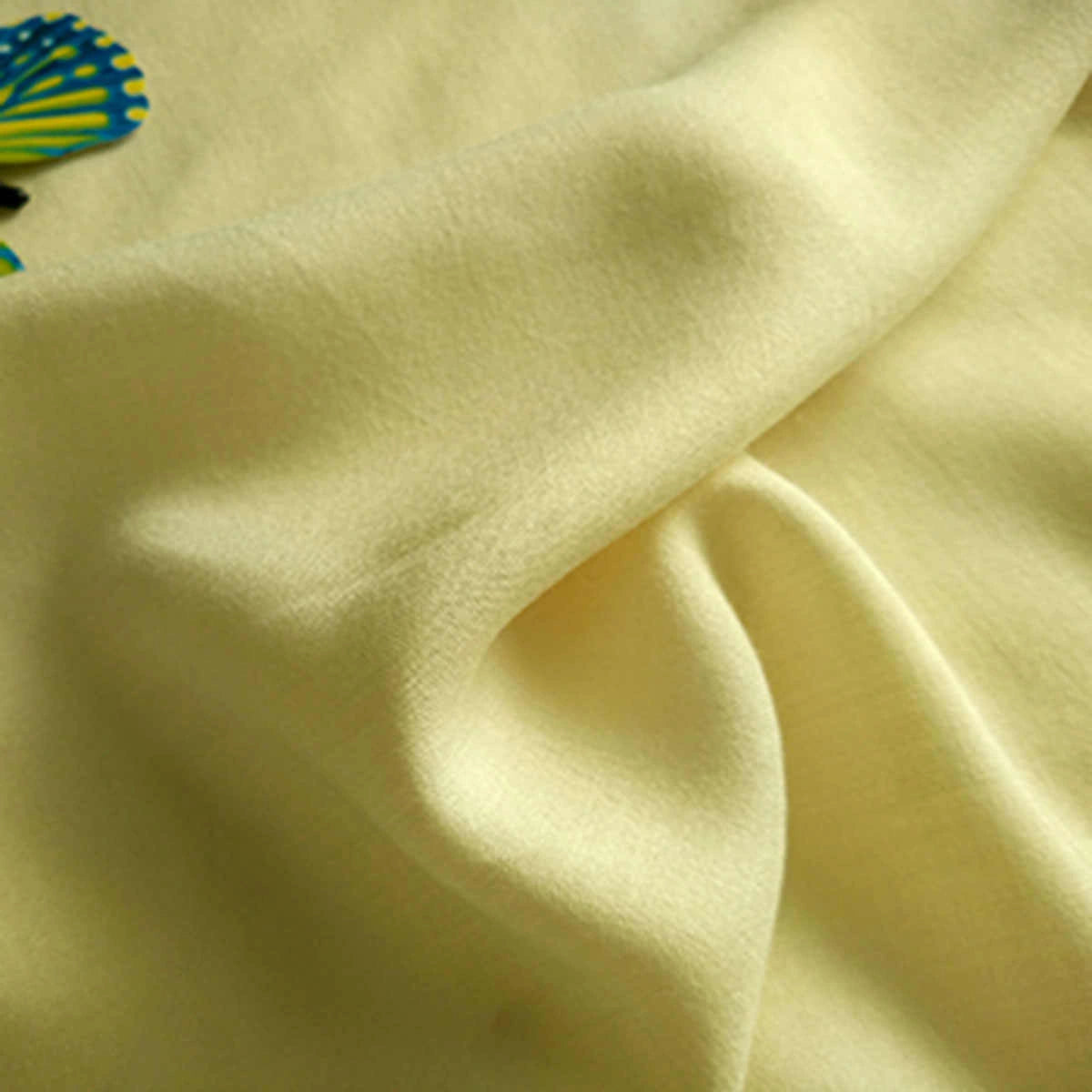 A close-up of Longan Craft’s Plain Color Jersey Viscose Fabric by the yard, 100% rayon viscose with a soft light yellow hue and smooth folds. A hint of a colorful object appears in the top left corner. Perfect fabric for dresses.