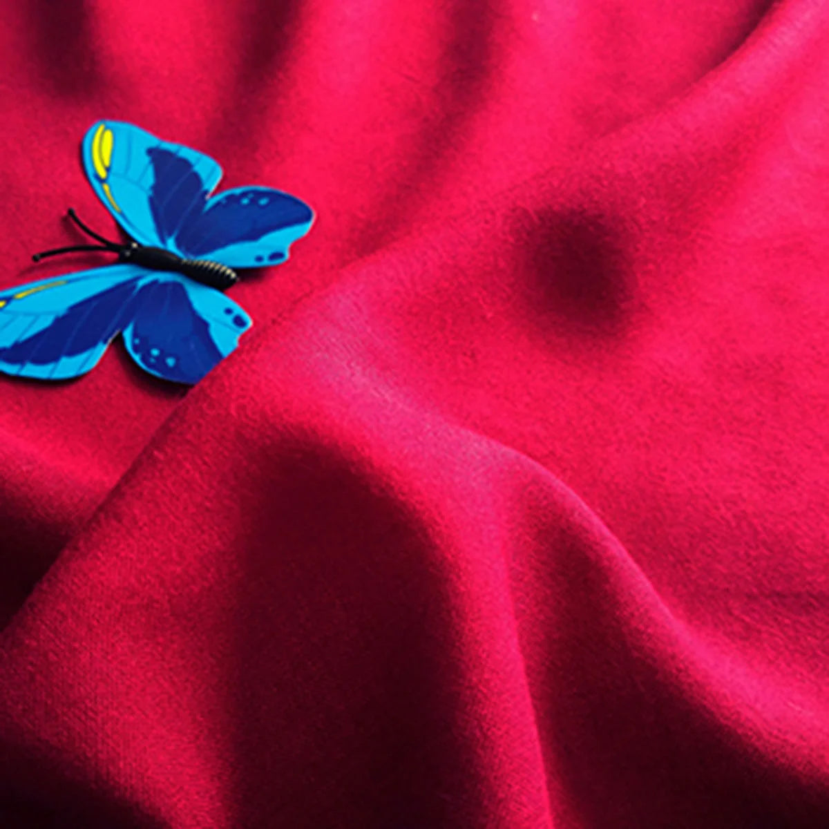 A vibrant blue butterfly rests on brightly colored red Plain Color Jersey Viscose Fabric By The Yard from Longan Craft, a soft 100% rayon viscose material perfect for dresses, with visible folds and gentle shadows.