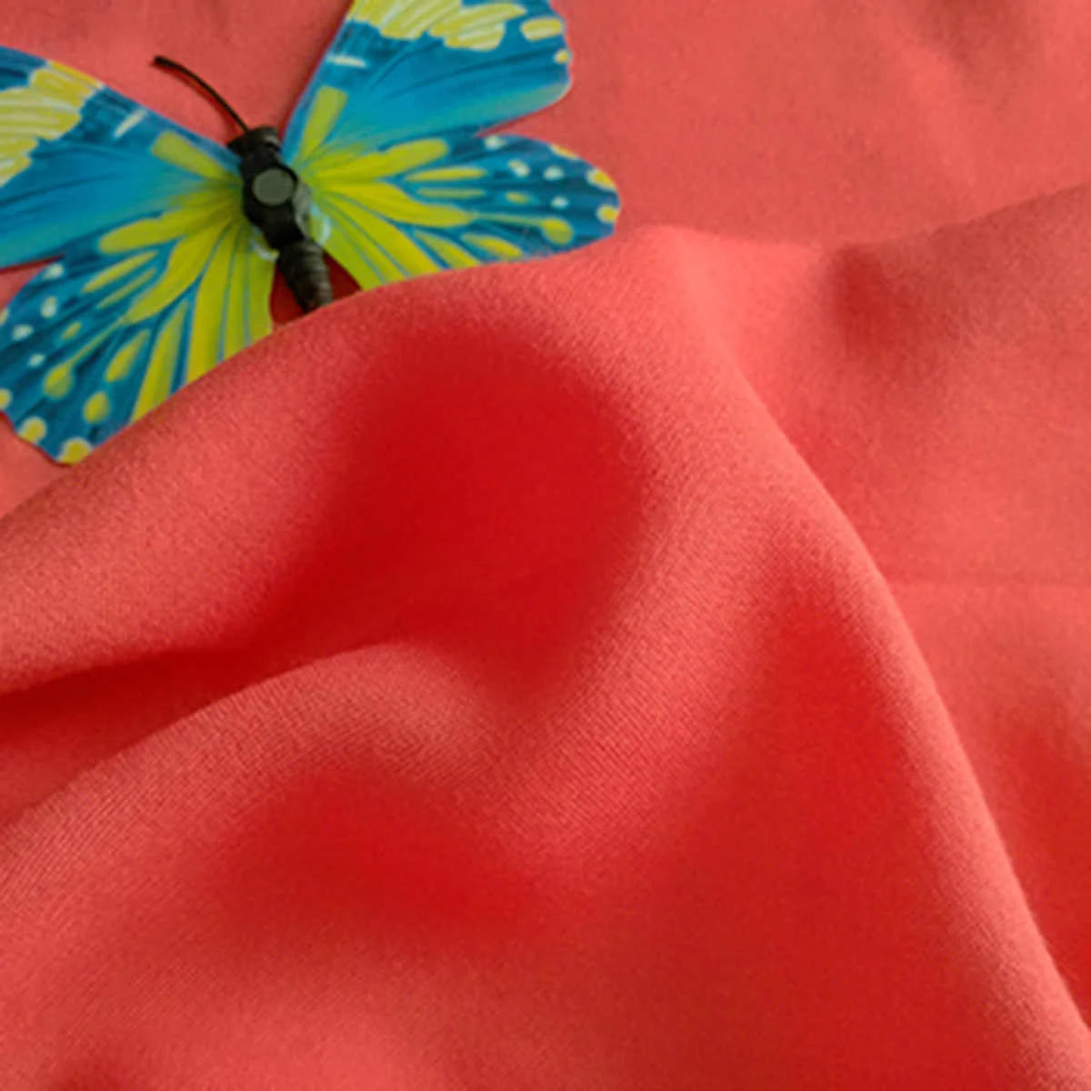 A vibrant blue and yellow butterfly rests on coral-colored Plain Color Jersey Viscose Fabric by Longan Craft, a soft 100% rayon viscose material perfect for dresses.