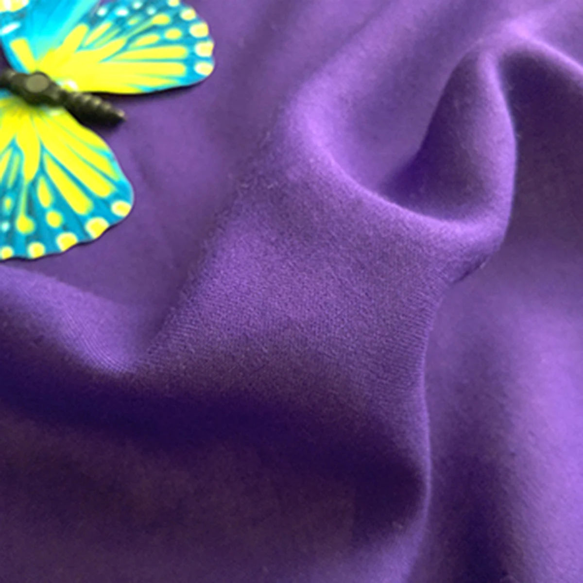 A close-up of a blue and yellow butterfly decoration resting on soft, purple Plain Color Jersey Viscose Fabric By The Yard by Longan Craft, made from 100% rayon viscose—perfect material for dresses with visible folds and texture.