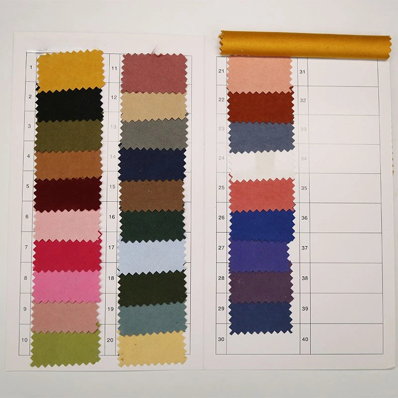 A fabric swatch card for Longan Craft's Multi Color Microsuede Polyester Peachskin Fabric By The Yard displays 26 rectangular samples in various colors, with handwritten numbers and a gold roll at the top right.