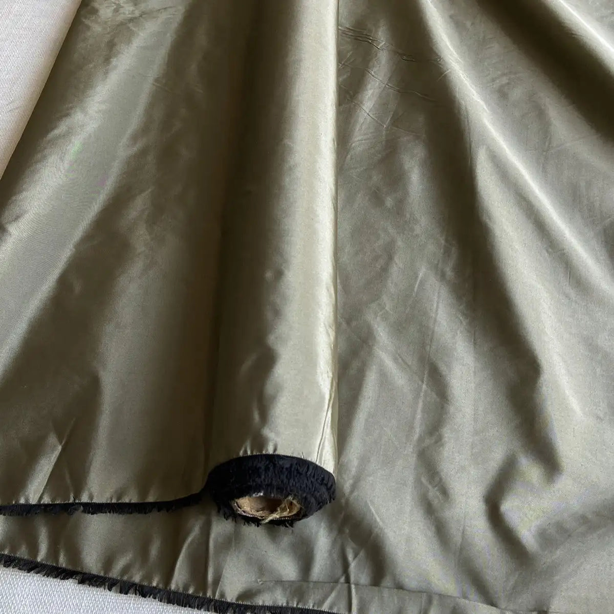 A close-up of Longan Craft’s olive-green Plain Color Taffeta Silk Fabric By The Yard, a stiff 100% mulberry silk bridal material, showing a shiny texture with black trim at the hem and a small frayed hole near the edge.