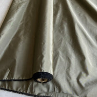 A close-up of Longan Craft’s olive-green Plain Color Taffeta Silk Fabric By The Yard, a stiff 100% mulberry silk bridal material, showing a shiny texture with black trim at the hem and a small frayed hole near the edge.