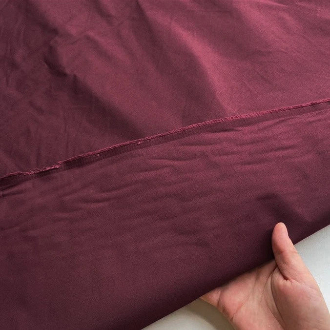 A hand lifts a piece of maroon Plain Color Taffeta Silk Fabric By The Yard from Longan Craft, showing its smooth, slightly shiny finish and stitched edge—a stiff, high quality 100% Mulberry silk perfect for bridal wear.