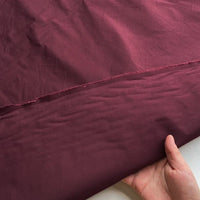 A hand lifts a piece of maroon Plain Color Taffeta Silk Fabric By The Yard from Longan Craft, showing its smooth, slightly shiny finish and stitched edge—a stiff, high quality 100% Mulberry silk perfect for bridal wear.