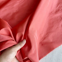 A hand holds and gathers Longan Craft's Plain Color Taffeta Silk Fabric by the yard—a stiff, high-quality 100% mulberry silk in coral pink—showing its elegant drape and texture, ideal for bridal wear, over a light surface.